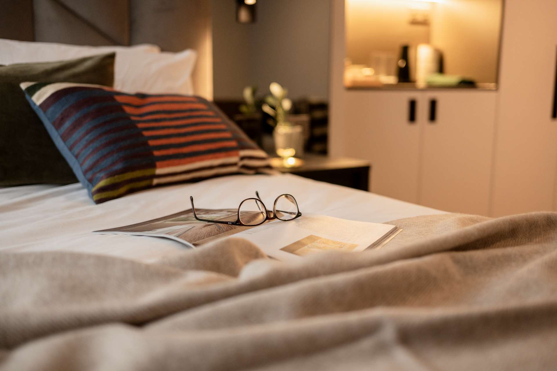 Modern apartment-style bedroom at The Hobson Cambridge by Adina with comfortable bed, soft lighting, reading glasses and magazine, ideal for long stays in Cambridge.
